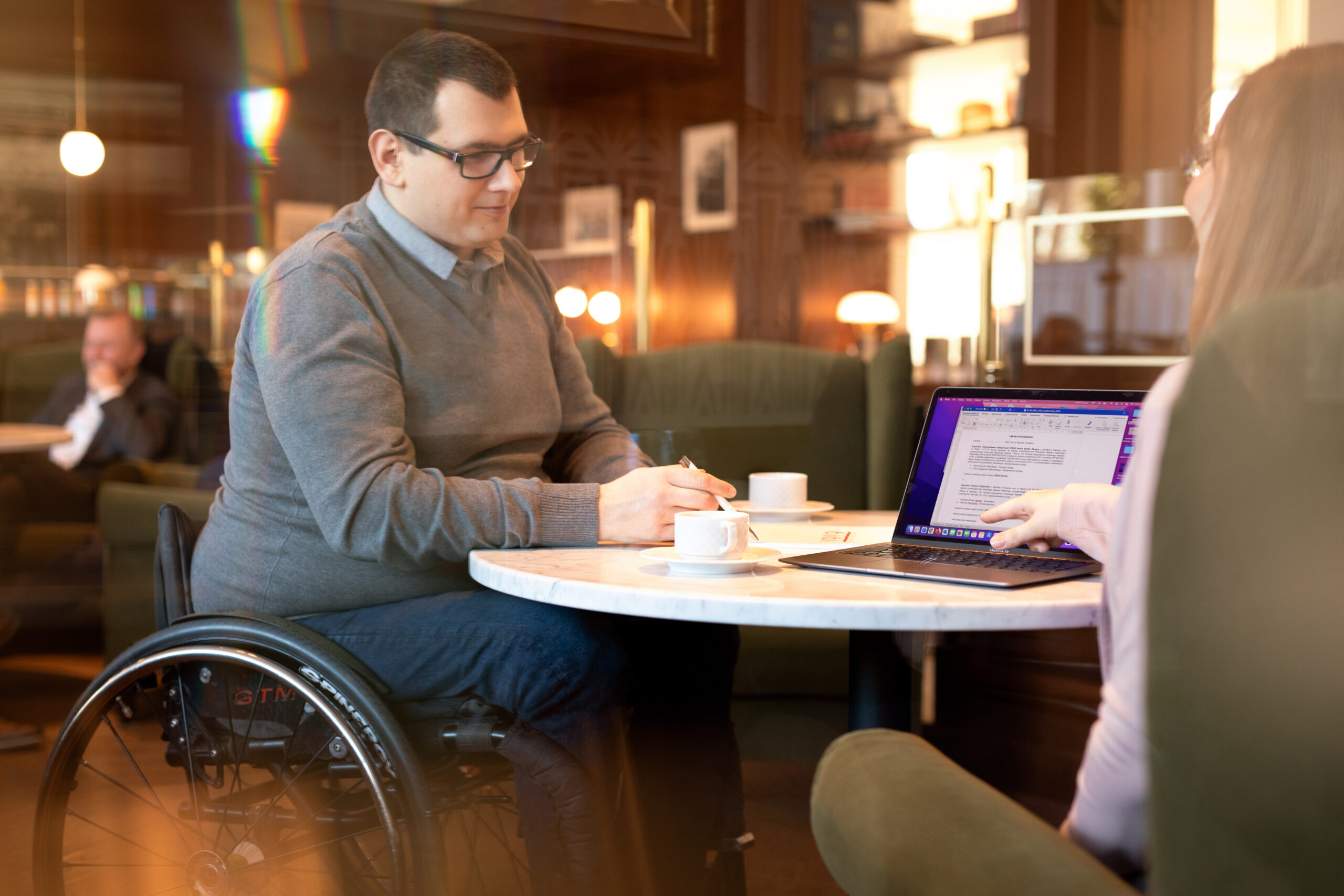 Mężczyzna poruszający się na wózku rozmawia w kawiarni z kobietą. Na stoliku stoją filiżanki i laptop. 